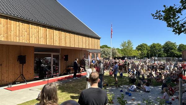 Et stort antal børn og voksne er samlet foran Stjær Børnehus til åbningsceremoni. Borgmesteren står ved en rød løber foran hovedindgangen, mens publikum sidder og står i solen.